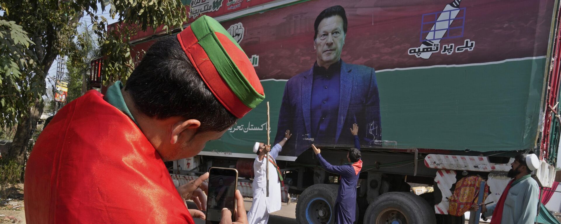 A supporter of former Prime Minister Imran Khan takes photos with mobile phone to his colleagues with huge Khan's poster painted on a truck neat the Khan's residence, in Lahore, Pakistan, Thursday, March 16, 2023. - Sputnik India, 1920, 27.04.2023