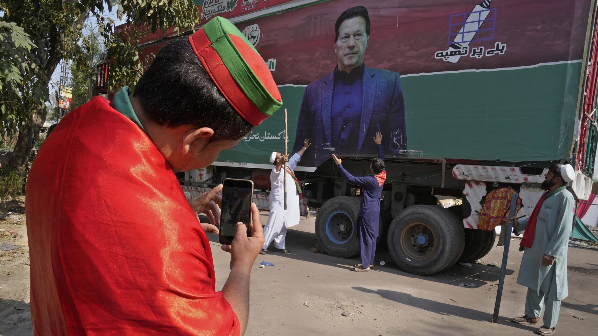 A supporter of former Prime Minister Imran Khan takes photos with mobile phone to his colleagues with huge Khan's poster painted on a truck neat the Khan's residence, in Lahore, Pakistan, Thursday, March 16, 2023. - Sputnik India, 1920, 17.03.2023
