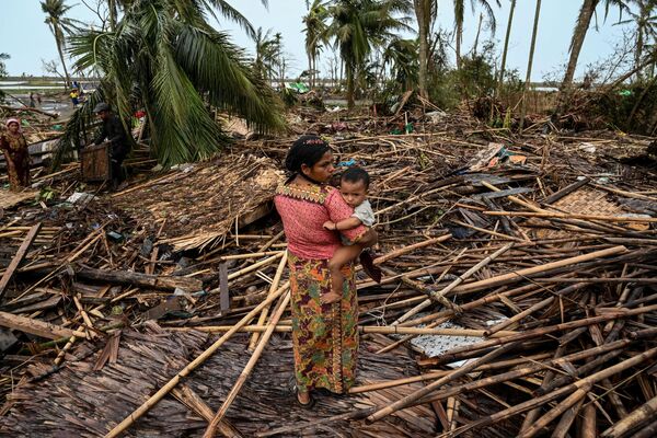16 मई, 2023 को सितवे में बसारा शरणार्थी शिविर में अपने नष्ट हुए घर के बगल में एक रोहिंग्या महिला अपने बच्चे को ले जा रही है(Photo by SAI Aung MAIN / AFP) - Sputnik भारत