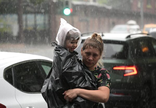 रूस के मास्को में भारी बारिश के दौरान सड़क पर रेन जैकेट पहने हुए एक बच्चे को ले जाती एक महिला। - Sputnik भारत
