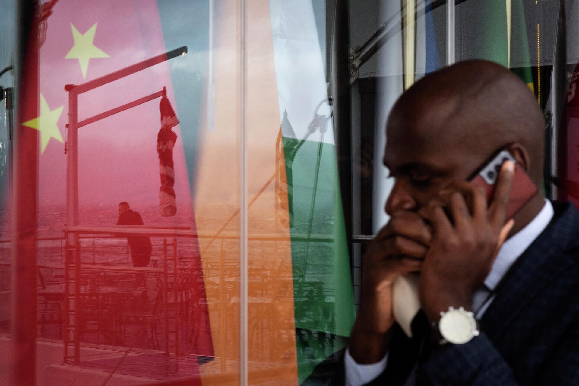 A man talks on a mobile phone as he walks past a view of a reflection on a window of another man staring at the sea in front of the national flags of the on the the BRICS (India, Russia, China, South Africa, Brazil) countries during the BRICS Foreign Ministers Meeting in Cape Town, on June 02, 2023. - Sputnik भारत, 1920, 19.08.2023