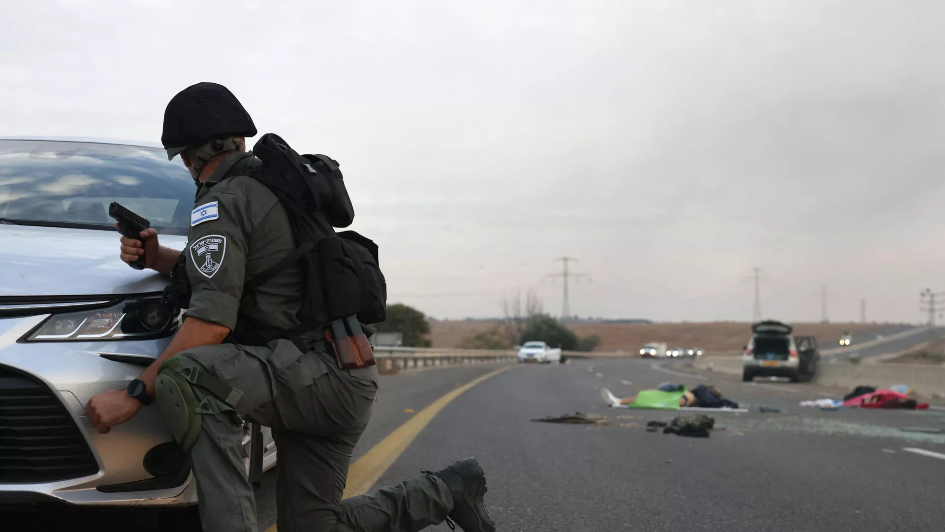 An Israeli soldier take cover behind a car as he looks toward bodies on a main road near the Gevim Kibbutz, close to the border with Gaza on October 7, 2023.