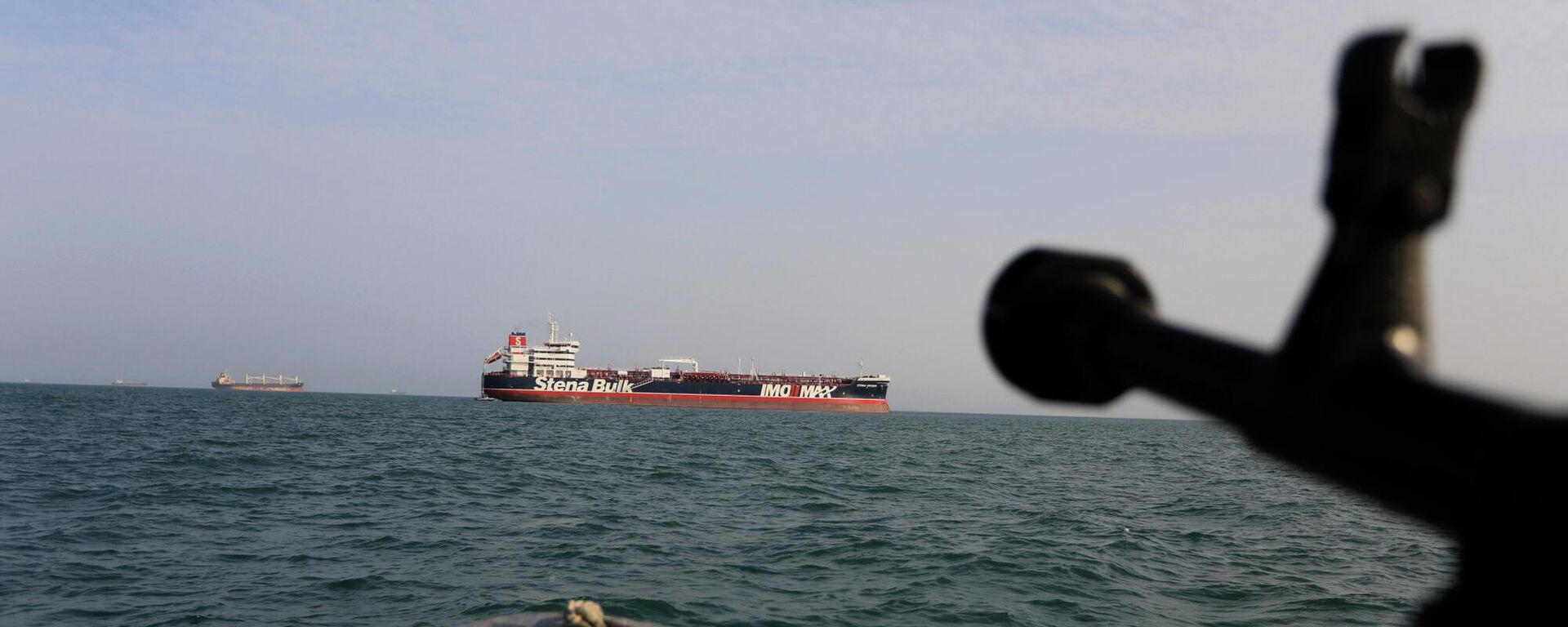 In this Sunday, July 21, 2019 photo, a speedboat of Iran's Revolutionary Guard trains a weapon toward the British-flagged oil tanker Stena Impero, which was seized in the Strait of Hormuz on Friday by the Guard, in the Iranian port of Bandar Abbas. Global stock markets were subdued Monday while the price of oil climbed as tensions in the Persian Gulf escalated after Iran's seizure of a British oil tanker on Friday.  - Sputnik India, 1920, 24.03.2026