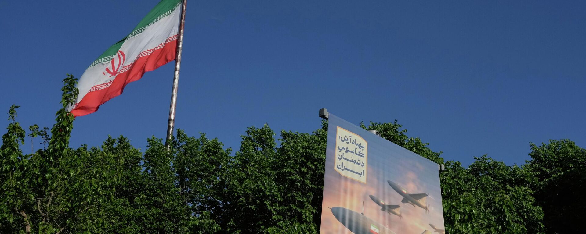 A banner shows a graphic depicting Iranian-built drones with a sign in Farsi that reads: Arash drone is the nightmare for Iran's enemies, as an Iranian flag waves at left, in Tehran, Iran, Tuesday, April 21, 2026.  - Sputnik भारत, 1920, 19.05.2026